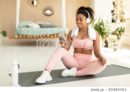 Positive fit black woman with towel on shoulders and wireless headphones sitting on fitness mat and listening to music Positive fit black woman with towel on shoulders and wireless headphones sitting on fitness mat and listening to music 93984561