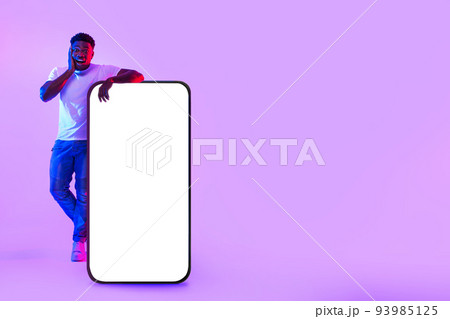 Surprised young black man standing near giant cellphone with empty white screen in neon light, mockup for mobile app Surprised young black man standing near giant cellphone with empty white screen in neon light, mockup for mobile app 93985125