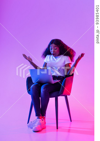 Full length of shocked young black woman using laptop, sitting in armchair, opening mouth in surprise in neon light 93985401