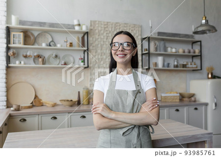 Portrait of a young beautiful woman cook, confectioner. She is standing in a beige apron and glasses in the kitchen, her arms crossed. He looks at the camera and smiles. 93985761