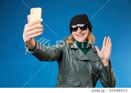 Young friendly fun woman 30s wearing stylish green jacket doing selfie shot on mobile cell phone talking by video call waving hand greeting isolated on blue background studio portrait. Young friendly fun woman 30s wearing stylish green jacket doing selfie shot on mobile cell phone talking by video call waving hand greeting isolated on blue background studio portrait. 93986052