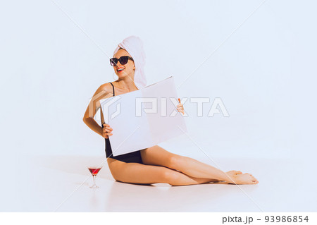 Portrait of beautiful young girl in black swimming suit relaxing, reading magazine, posing over grey background 93986854