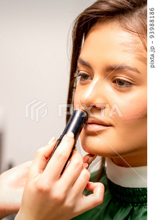 Beautiful young brunette woman receiving makeup with stick concealer on her face in a beauty salon 93988186