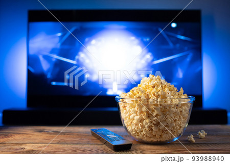 A glass bowl of popcorn and remote control in the background the TV works. Evening cozy watching a movie or TV series at home A glass bowl of popcorn and remote control in the background the TV works. Evening cozy watching a movie or TV series at home 93988940