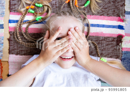 Close up portrait of caucasian cheerful girl with pigtails lying on the floor on a woven striped rug, smiling, playing hide and seek, covering her eyes with her hands. Digital detox, lifestyle. Close up portrait of caucasian cheerful girl with pigtails lying on the floor on a woven striped rug, smiling, playing hide and seek, covering her eyes with her hands. Digital detox, lifestyle. 93991805