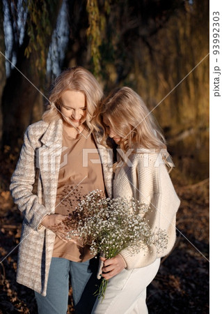 Adult daughter hugs her pregnant mom on a sunny autumn day in nature. Blonde woman holding flowers in her hand. Women wearing beige clothes. 93992303