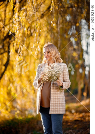 Portrait of middle-aged pregnant woman outdoors at park. Middle age pregnant woman expecting baby at aged pregnancy. Blonde woman wearing brown sweater and beige coat. 93992346