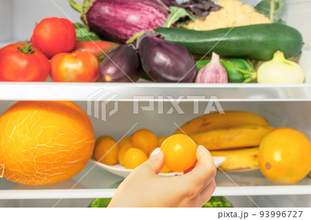 woman's hand takes a fruit from the refrigerator. The refrigerator is filled with vegetables and fruits. Weight loss diet concept. woman's hand takes a fruit from the refrigerator. The refrigerator is filled with vegetables and fruits. Weight loss diet concept. 93996727