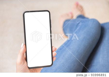 Mockup image of woman's hands holding mobile phone with blank screen. Woman is sitting on the sofa with her legs stretched out. 93996891