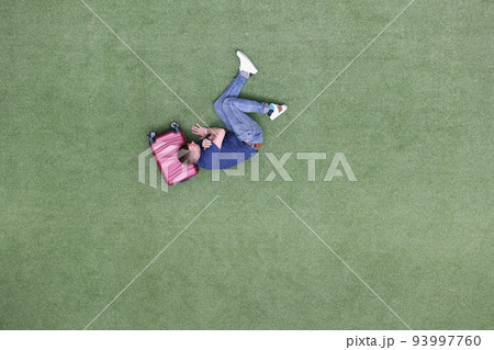 Young man lies on suitcase on green lawn Young man lies on suitcase on green lawn 93997760