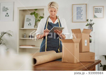 Packing parcel or package in shop, online fashion order on tablet, clothes boutique or design workshop. Mature tailor or seamstress packaging, boxing or wrapping clothing product for courier delivery 93999984