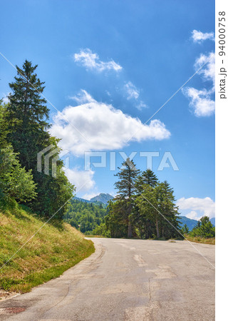 Curvy open road or street through green tall trees with a cloudy blue sky. Landscape view of a path in a scenic and peaceful location surrounded by nature or lush foliage on a summer day Curvy open road or street through green tall trees with a cloudy blue sky. Landscape view of a path in a scenic and peaceful location surrounded by nature or lush foliage on a summer day 94000758