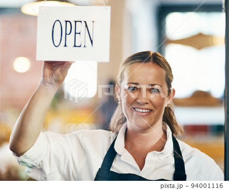 Cafe owner opening her new startup, store or welcoming customers to a restaurant. Caucasian manager reopening her business after closing down. Waitress holding open sign at window or entrance. Cafe owner opening her new startup, store or welcoming customers to a restaurant. Caucasian manager reopening her business after closing down. Waitress holding open sign at window or entrance. 94001116