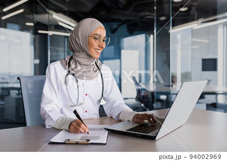 Arab muslim woman in hijab doctor working in modern clinic office with laptop, doctor on paper work in white medical coat with stethoscope and glasses. Arab muslim woman in hijab doctor working in modern clinic office with laptop, doctor on paper work in white medical coat with stethoscope and glasses. 94002969