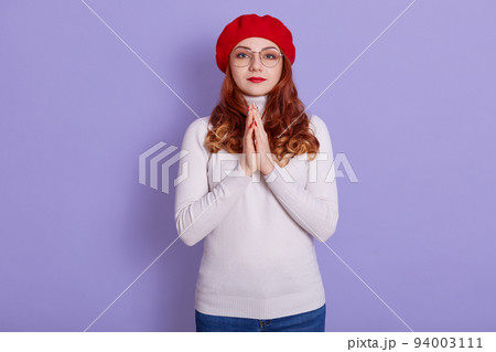 Close-up portrait of nice attractive pretty girl keeping arms together asking favor, expecting luck isolated over lilac pastel background, female wearing red beret, winsome woman with wavy hair. Close-up portrait of nice attractive pretty girl keeping arms together asking favor, expecting luck isolated over lilac pastel background, female wearing red beret, winsome woman with wavy hair. 94003111