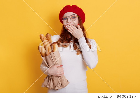 Young beautiful red haired woman wearing french beret, holding bread, covering mouth with hand, has surprised expression, looks at camera with pleasant smile, isolated over yellow background. Young beautiful red haired woman wearing french beret, holding bread, covering mouth with hand, has surprised expression, looks at camera with pleasant smile, isolated over yellow background. 94003137