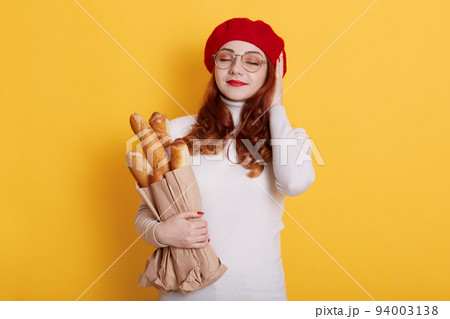 Pretty young redhead woman stands with closed eyes and pleasant smile. Glad ginger girl feels happy isolated on yellow wall, holding long bread baguettes, wearing red beret and white shirt. Pretty young redhead woman stands with closed eyes and pleasant smile. Glad ginger girl feels happy isolated on yellow wall, holding long bread baguettes, wearing red beret and white shirt. 94003138