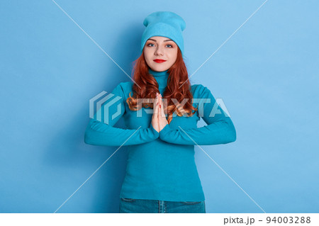 Lovely female makes request, keeps palms presses together, asks for something in close person, has calm expression, dressed casually, isolated over blue background. Pleading woman in cap and sweater. Lovely female makes request, keeps palms presses together, asks for something in close person, has calm expression, dressed casually, isolated over blue background. Pleading woman in cap and sweater. 94003288