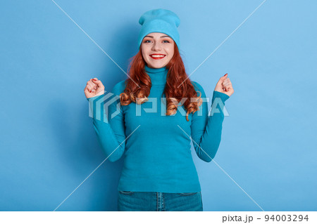 Happy laughing woman with red wavy hair, looking directly at camera with toothy smile, clenches fists like winner, rejoices her success, wearing casual attire. 94003294