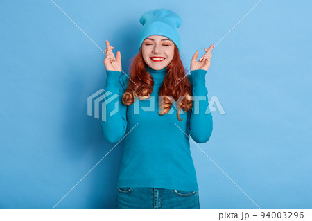 Portrait of happy young girl with red wavy hair, keeps fingers crossed and eyes closed, wearing blue sweater and cap, wishes best, expressing positive emotions. 94003296