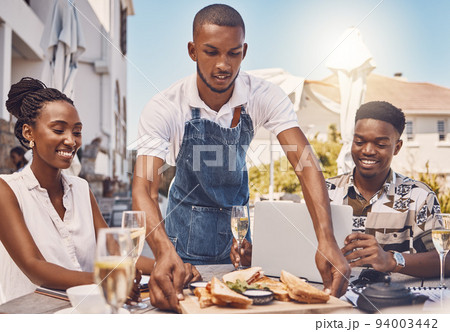 Waiter serving food to couple at a restaurant, worker giving service and food on table at a cafe in summer. Happy man and woman eating lunch at a shop, happy with server and date at coffee shop 94003442