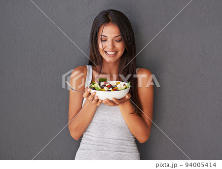 Healthy young female eating her fresh food salad bowl. Smiling beautiful woman holding and enjoying eating her clean green diet dish of vegetables as part of her vegan wellness lifestyle 94005414