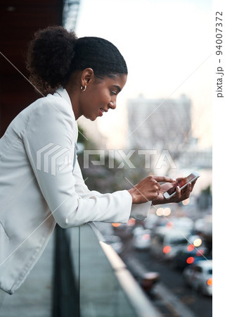 Relaxed business woman texting on her phone while standing on a balcony on her lunch break. Young corporate female browsing the internet or scrolling on social media after work in a city 94007372