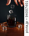 The barman throws ice cubes into a cocktail glass. The splashes of the drink are beautifully scattered on a wooden tray. 94007401