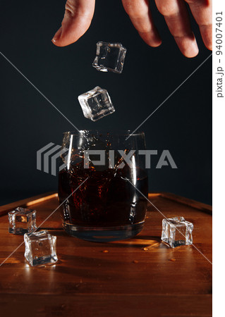 The barman throws ice cubes into a cocktail glass. The splashes of the drink are beautifully scattered on a wooden tray. 94007401