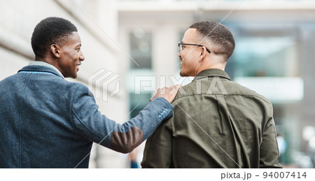 Two young businessmen smiling, talking and walking through the city together, excited for their new project. Happy multiracial coworkers strolling with his hand on his friends shoulder while laughing 94007414