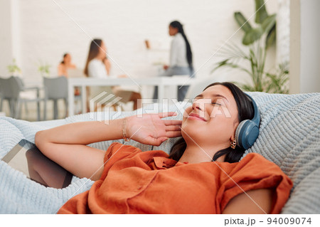 Creative wearing headphones, listening to music from tablet and relaxing, feeling inspired or motivated in a casual workspace. Happy and zen business woman lying on office bean bag and taking a break 94009074