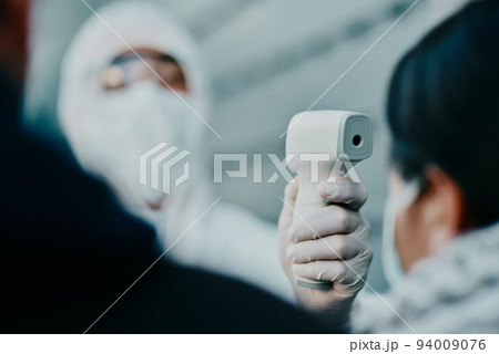 Medical healthcare worker, scanning temperature of covid patient for safety against virus pandemic, wearing protective suit. Doctor in hazmat suit checking with thermometer for potential outbreak. 94009076