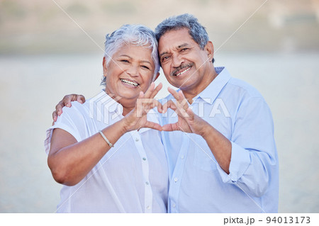 A happy senior couple with a heart sign with their fingers and enjoying fresh nature air on vacation at beach while bonding. Portrait of retired couple hugging at beach with smile and love together 94013173