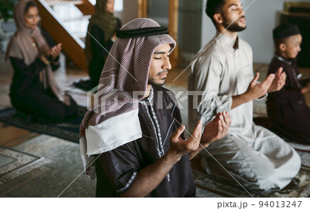 Young muslim family pray together, sit on knees at home practicing Islam. Religious women wearing hijab and spiritual men wear traditional attire, close eyes and concentrate on prayer with hands up. 94013247