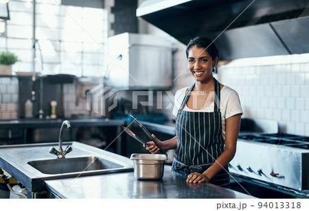 Cooking, making food and working as a chef in a commercial kitchen with tongs and industrial equipment. Portrait of a female cook preparing a meal for lunch, dinner or supper in a restaurant or cafe 94013318