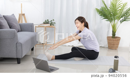 Beautiful Asian woman in sportswear is warming up or doing light exercises in the house with dumbbells and a protein shake or bottle of water and using a laptop at home, Healthy exercise. 94020556