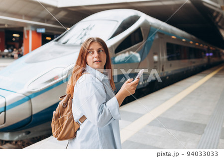 Young redhhead woman waiting train on station platform with backpack and using smart phone. Railroad transport concept, Traveler. Woman traveler walking at train station Young redhhead woman waiting train on station platform with backpack and using smart phone. Railroad transport concept, Traveler. Woman traveler walking at train station 94033033