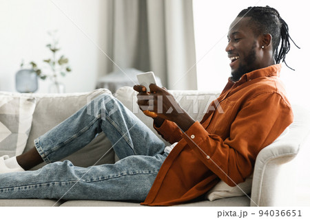 Joyful african american man reclining on couch at home, chatting with friends or playing games on cellphone, copy space Joyful african american man reclining on couch at home, chatting with friends or playing games on cellphone, copy space 94036651