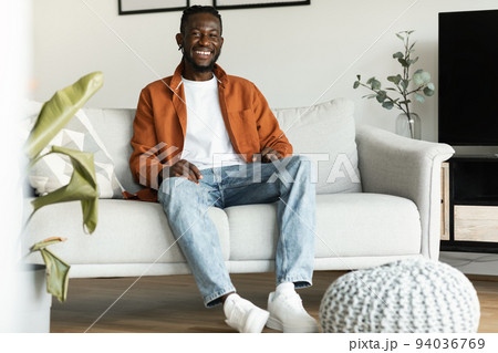 Positive black man sitting on sofa at home, relaxing alone at weekend and smiling at camera, free space Positive black man sitting on sofa at home, relaxing alone at weekend and smiling at camera, free space 94036769