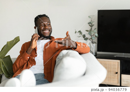 Making call. Happy african american man talking on smartphone and gesturing, resting on sofa at home, free space 94036779