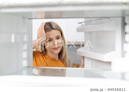 Sad blonde woman looking inside open empty fridge Sad blonde woman looking inside open empty fridge 94036811