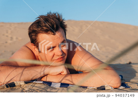 Looking away young 32 years old Caucasian man lies on striped towel and sunbathes on sandy beach on sand dune. Close-up summer headshot lifestyle three quarter portrait. Suntan, resting, relaxing. 94037149