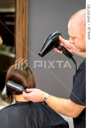 Male hairdresser drying short hair of young caucasian brunette woman with a black hairdryer and black round brush in a hairdresser salon 94039782