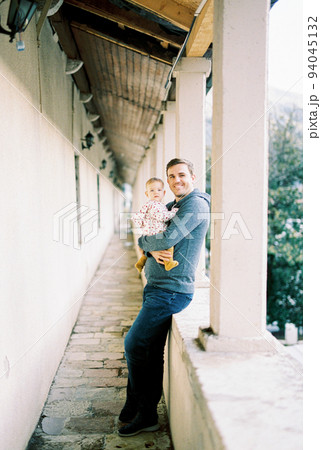 Smiling dad with a baby in his arms stands on the terrace leaning against a stone fence 94045132