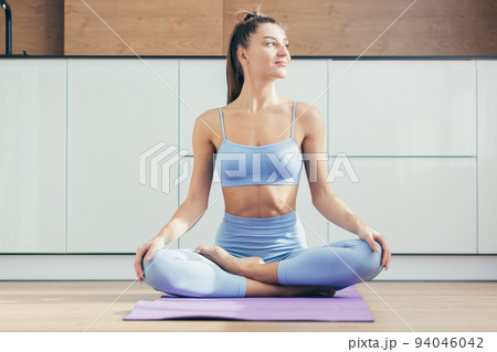 Yoga time. Portrait of a young beautiful woman practicing yoga at home, sitting on a mat in the lotus position, looking at the camera, smiling, relaxed 94046042