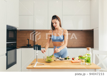 Young beautiful woman nutritionist teaches to cook online the Internet video call smartphone in the kitchen at home dish of fresh vegetable salad, looking at the camera, smiling Young beautiful woman nutritionist teaches to cook online the Internet video call smartphone in the kitchen at home dish of fresh vegetable salad, looking at the camera, smiling 94047233