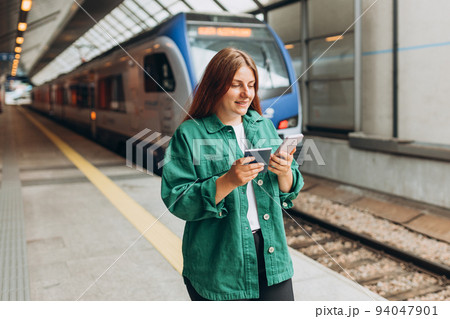 Young redhhead woman with passport waiting on station platform and using smart phone. Railroad transport concept. Travel to vacation by fast train. 94047901
