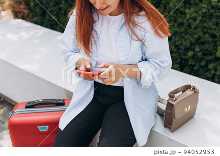 Woman with a red suitcase using smartphone in the European city. Hipster girl browsing Internet on a phone, texting and communicating outdoors. Travel concept 94052953