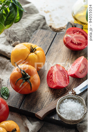 Raw tomatoes and knife on wooden cutting board surrounded by ingredients salt basil leaves and olive oil around. Rustic cooking background 94055034