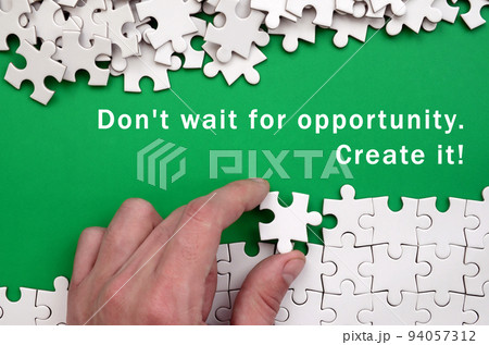 Don't wait for opportunity. Create it. The hand folds a white jigsaw puzzle and a pile of uncombed puzzle pieces lies against the background of the green surface 94057312